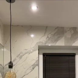 Ciemne żaluzje poziome w białej ramie okiennej na tle ściany z marmurowym wzorem, oświetlone lampą sufitową i wiszącą żarówką Edisona.