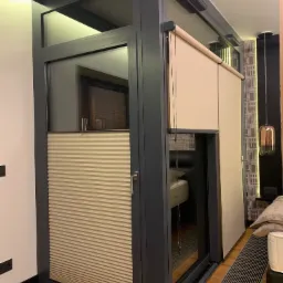 Beżowa plisa pozioma w oknie z szarą ramą, u góry roleta zwijana, widok na wnętrze sypialni z łóżkiem i lampą.