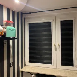 Pokój z oknem wyposażonym w rolety w stylu dzień i noc, z czarno-białymi pasami na ścianie i elementami dekoracyjnymi na półce.