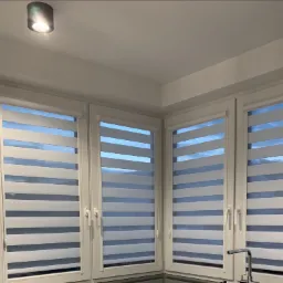 Trzy okna narożne z zamontowanymi roletami typu dzień i noc, widoczny fragment blatu kuchennego i bateria umywalkowa.