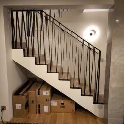 Vaanto - Nowoczesne schody z drewnianymi stopniami i czarną, geometryczną balustradą. Pod schodami kartony. Na ścianie okrągła lampa. Minimalistyczny design.