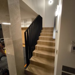 Vaanto - Klatka schodowa z drewnianymi stopniami i nowoczesną, czarną balustradą z metalu. Minimalistyczne oświetlenie, jasne ściany i marmurowe wykończenie kolumny.