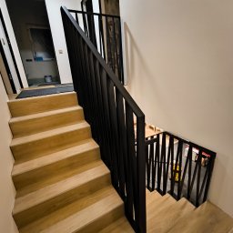 Vaanto - Drewniane schody z czarną, nowoczesną balustradą. Widok z góry, ukazujący konstrukcję i wykończenie. W tle widoczne pomieszczenia na różnych poziomach.