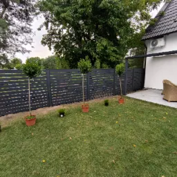 Nowoczesne ogrodzenie panelowe z poziomych desek w kolorze grafitowym. Na pierwszym planie trawnik z drzewkami w donicach. W tle dom z jasną elewacją i pergola.