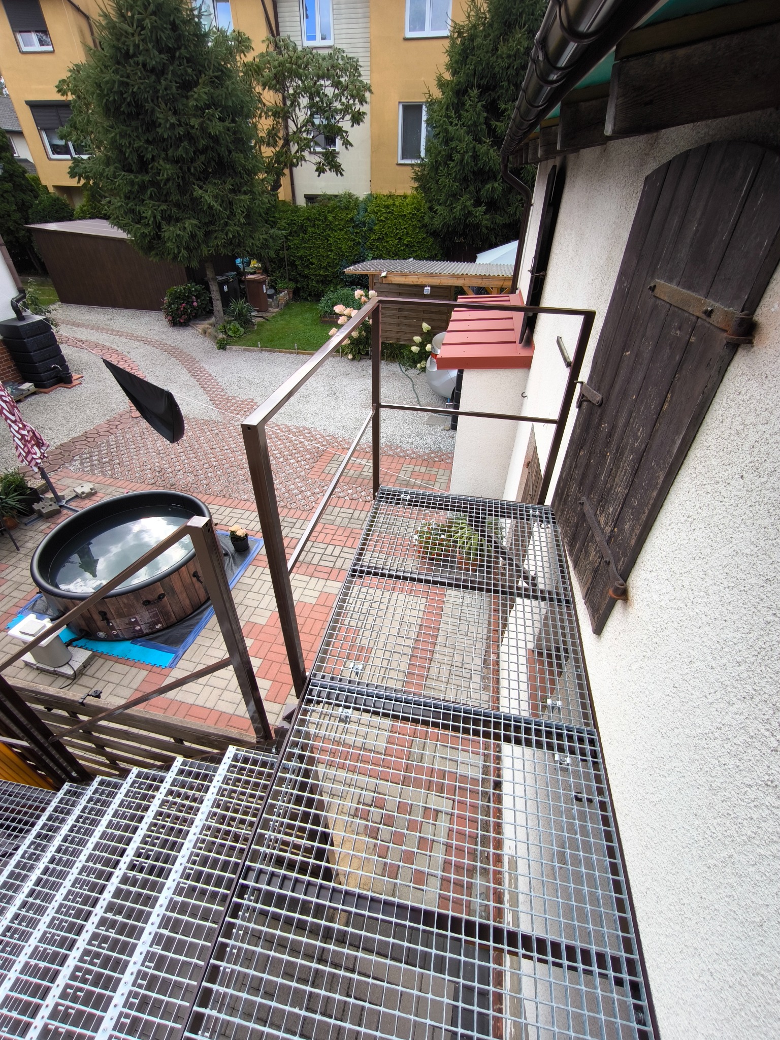 Metalowe schody kratowe prowadzą do podestu z balustradą. W tle budynek, ogród i jacuzzi. Widok z góry, z perspektywy osoby stojącej na schodach.