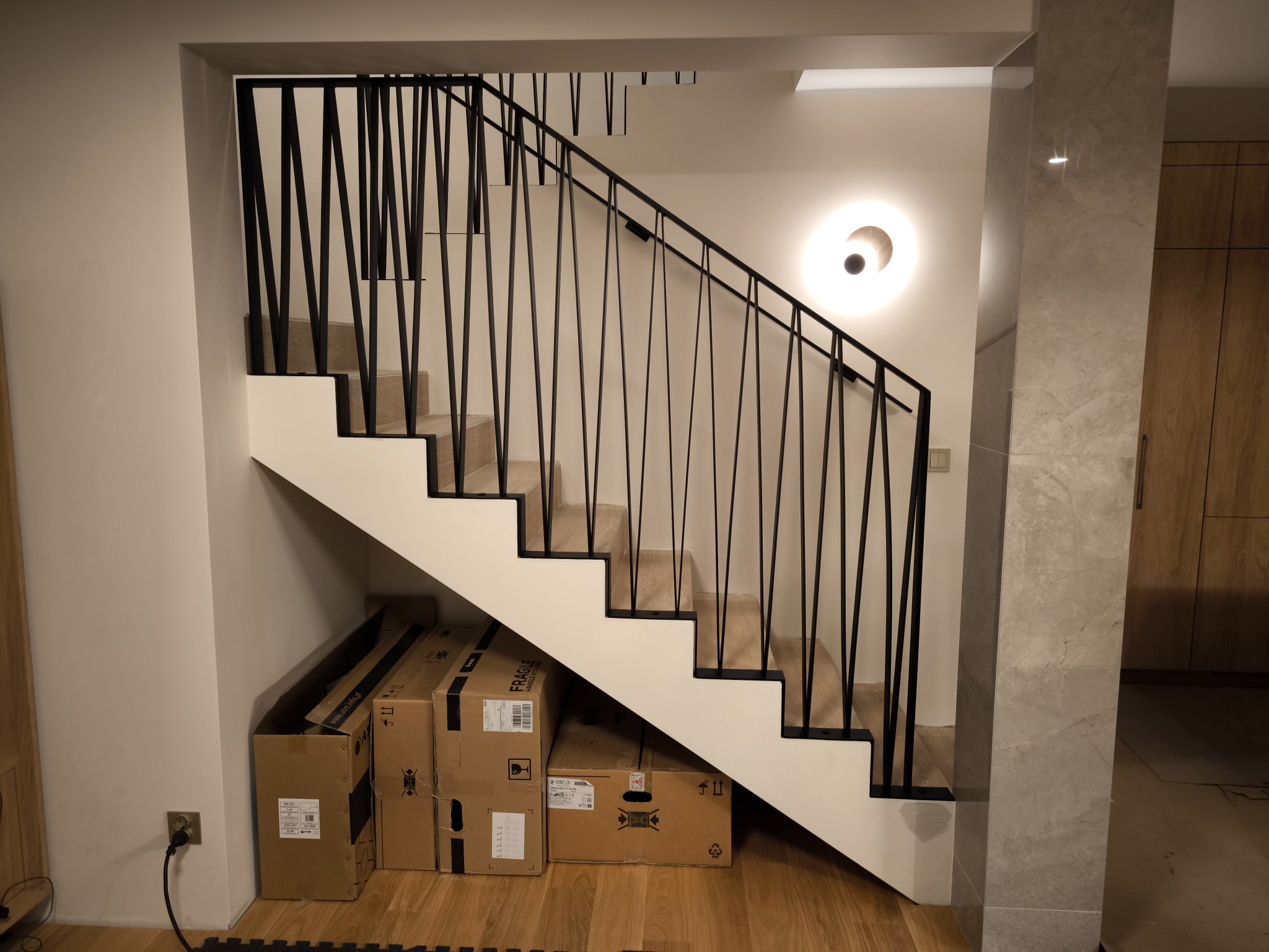Nowoczesne schody z drewnianymi stopniami i czarną, geometryczną balustradą. Pod schodami kartony. Na ścianie okrągła lampa. Minimalistyczny design.