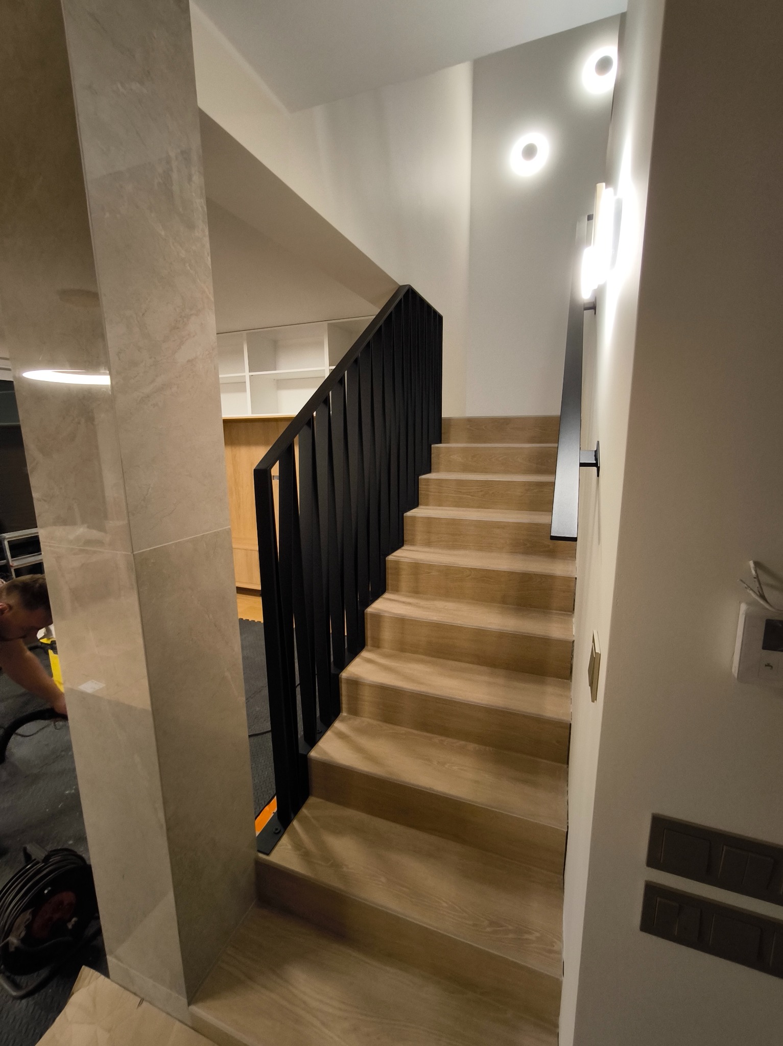 Klatka schodowa z drewnianymi stopniami i nowoczesną, czarną balustradą z metalu. Minimalistyczne oświetlenie, jasne ściany i marmurowe wykończenie kolumny.