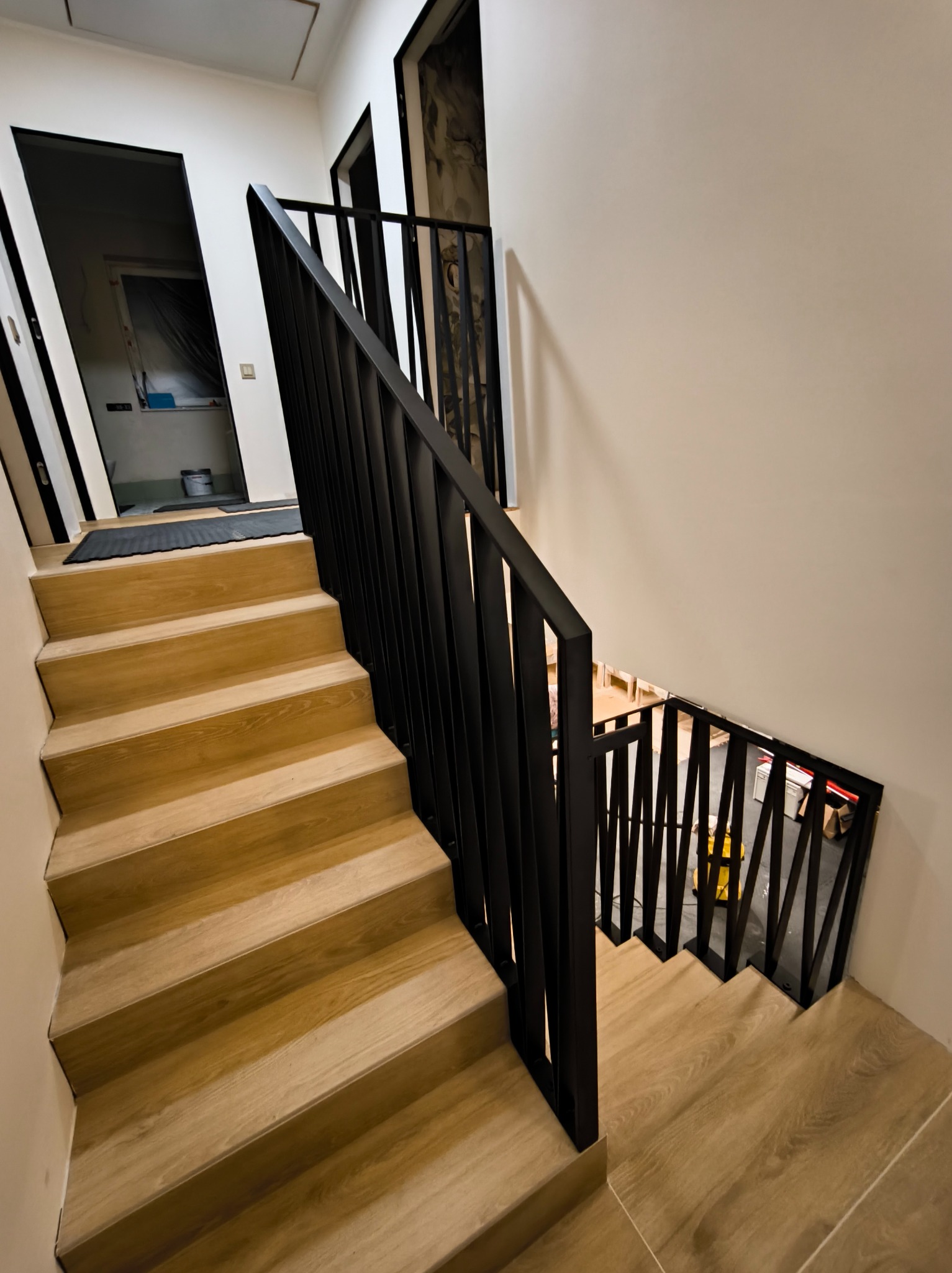 Drewniane schody z czarną, nowoczesną balustradą. Widok z góry, ukazujący konstrukcję i wykończenie. W tle widoczne pomieszczenia na różnych poziomach.