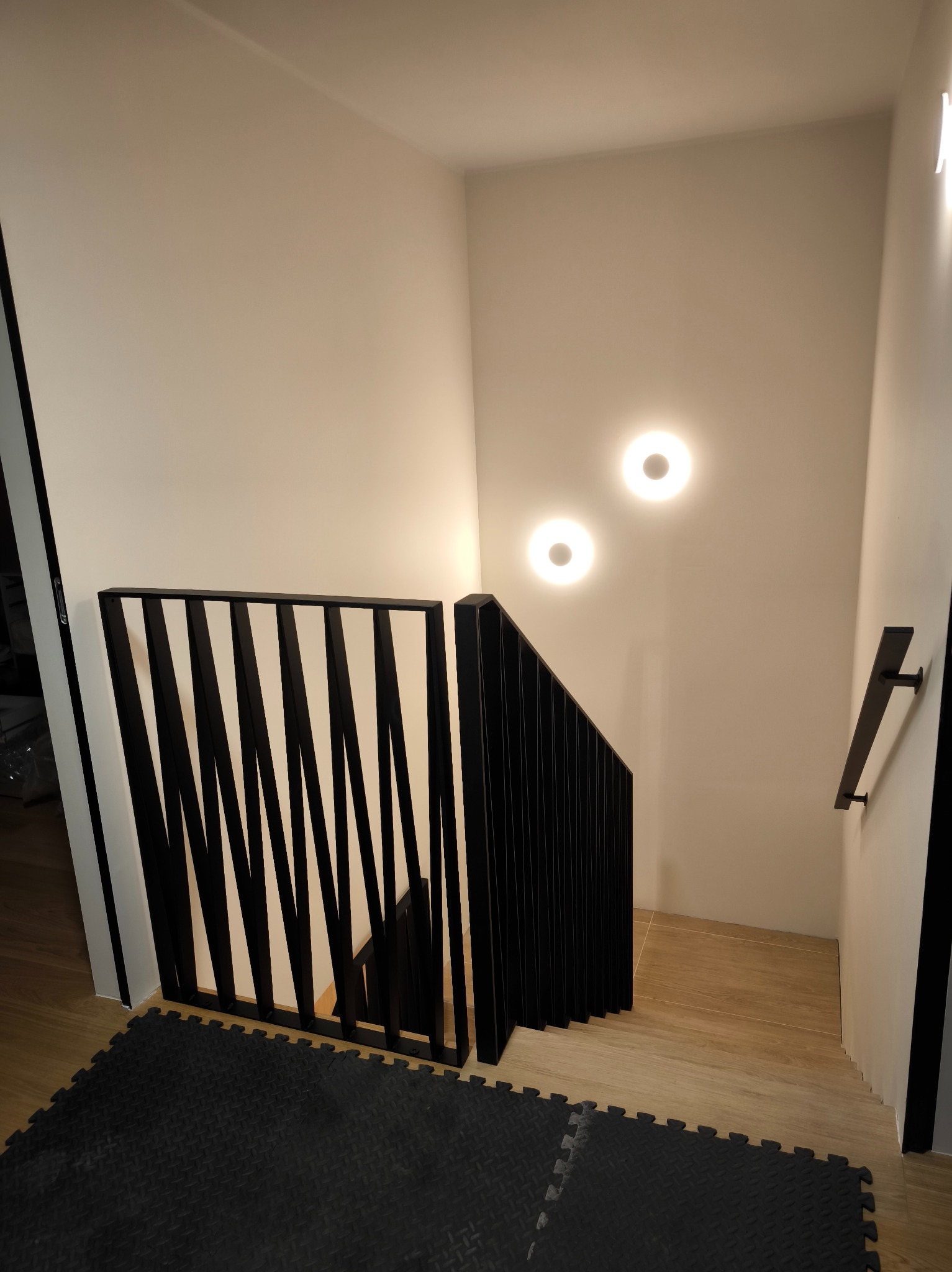 Nowoczesne schody z czarną balustradą o geometrycznym wzorze, oświetlone dwoma okrągłymi lampami, z widocznym fragmentem poręczy na ścianie.