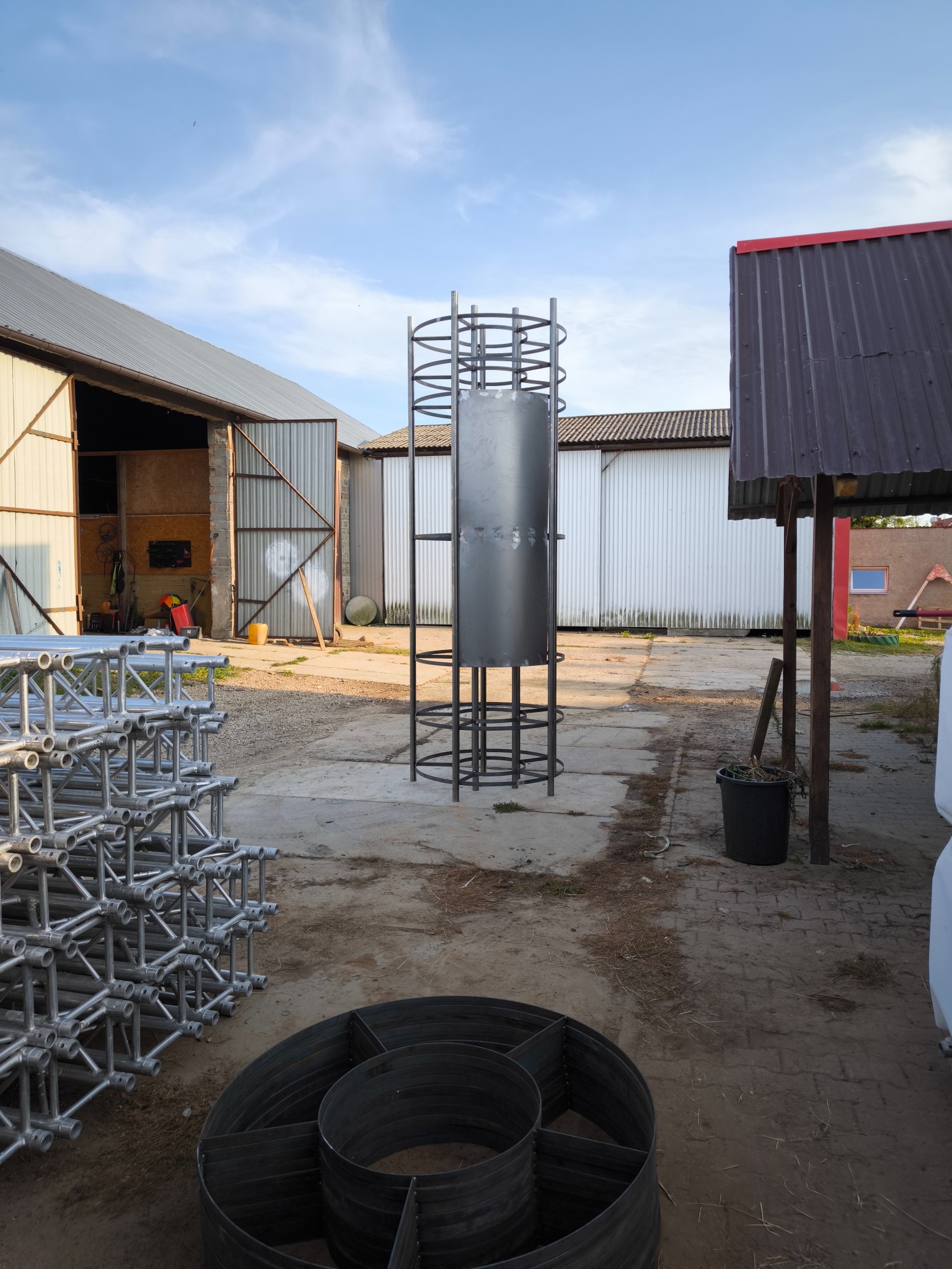 Konstrukcja stalowa z metalowym zbiornikiem w centrum kadru, otoczona elementami rusztowań i fragmentami metalowych pierścieni na zewnątrz, na tle zabudowań gospodarczych.