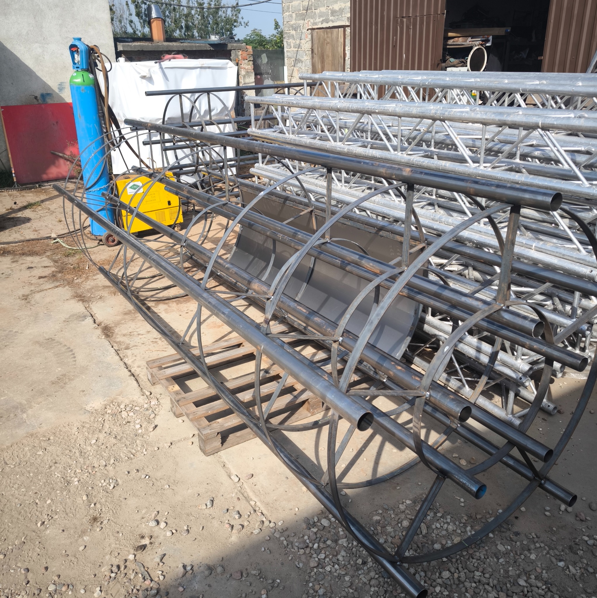 Konstrukcja z metalowych rur o okrągłym przekroju, spawana, z widocznym spawem, leżąca na ziemi obok butli z gazem i spawarki, z fragmentem budynku w tle.