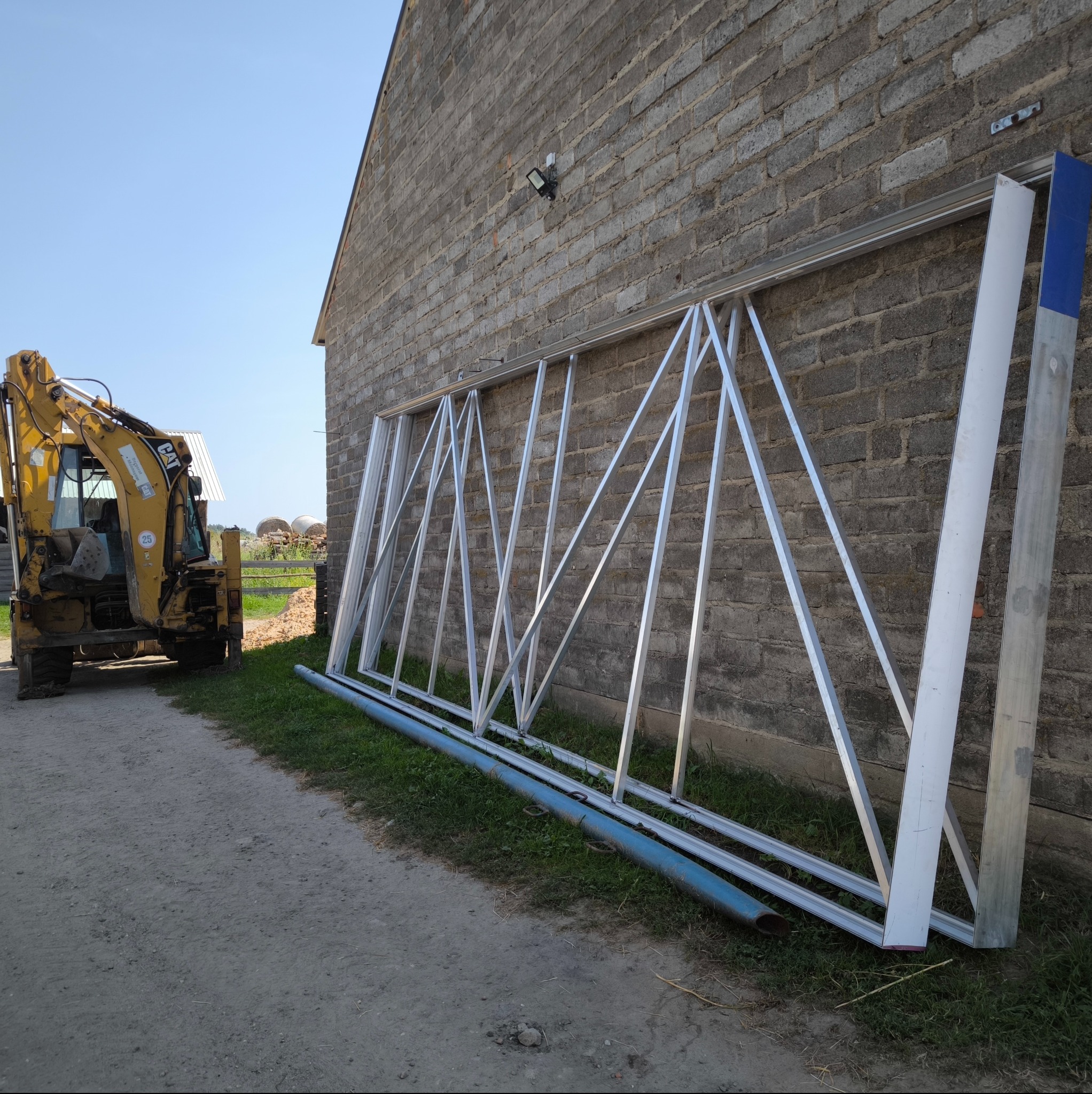 Konstrukcja metalowa z trójkątnymi wzmocnieniami oparta o budynek z cegły, obok stoi żółta koparka. Elementy przygotowane do montażu.
