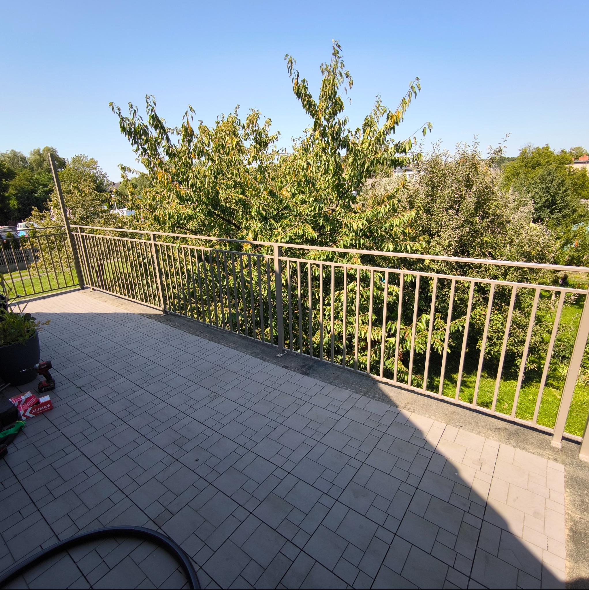 Beżowa, metalowa barierka balkonowa z widokiem na zieleń drzew w słoneczny dzień. Na posadzce balkonu leżą narzędzia.