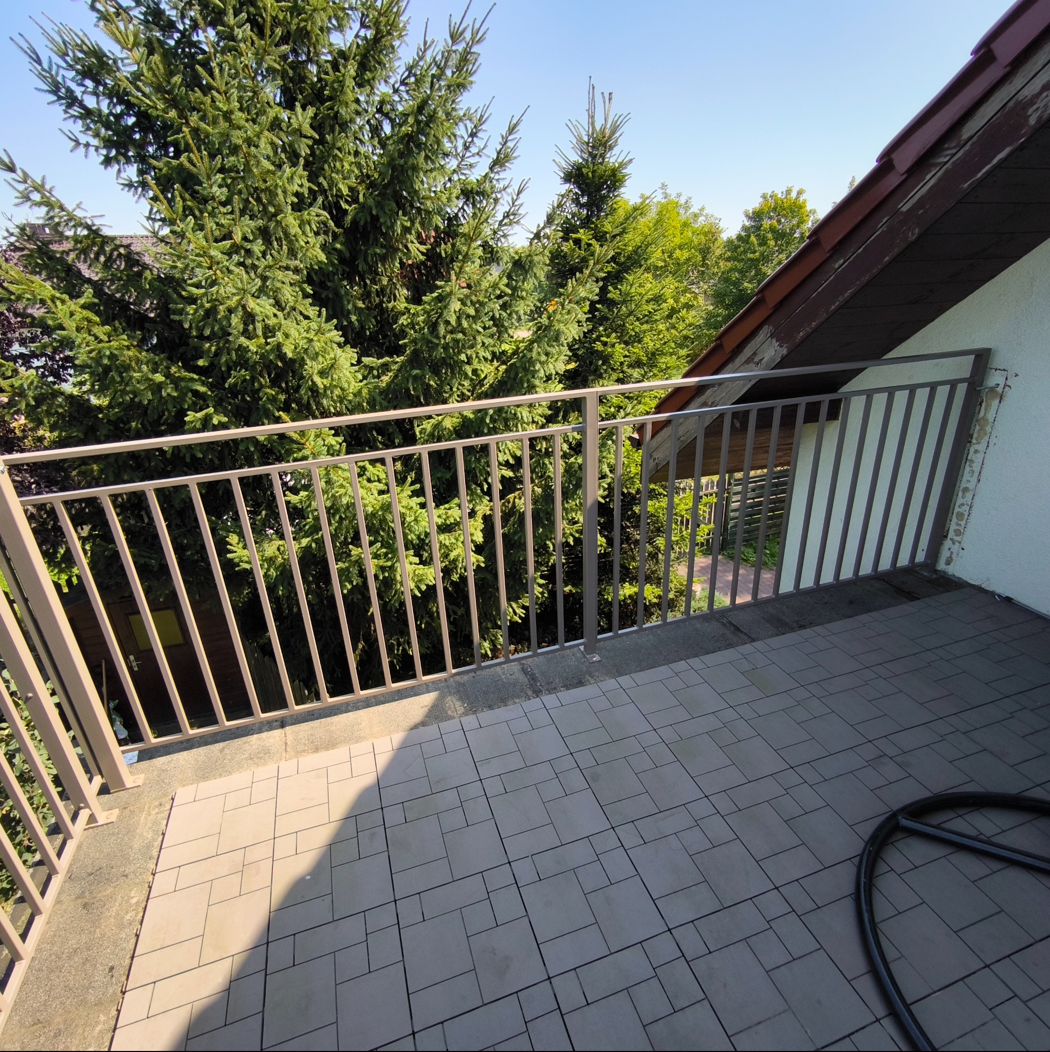Beżowa, metalowa balustrada balkonowa z pionowymi szczeblami, na tle zieleni drzew. Widok z góry na szare płytki balkonowe i fragment dachu.