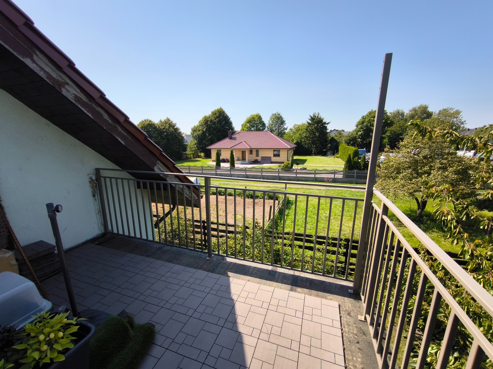 Balkon z widokiem na dom i ogród, zabezpieczony metalową balustradą w kolorze beżowym. Na podłodze balkonu ułożona szara, kwadratowa terakota.