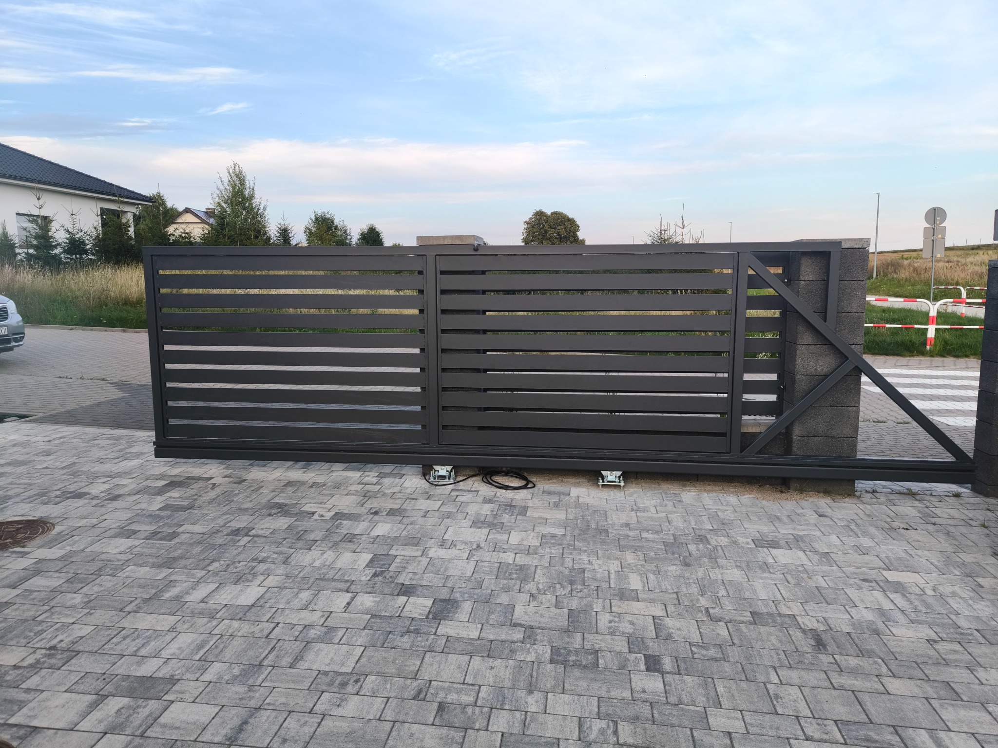 Nowoczesna, przesuwna brama wjazdowa w kolorze antracytowym z poziomymi panelami, zamontowana na kostce brukowej. Widoczne elementy konstrukcyjne i automatyka bramy.
