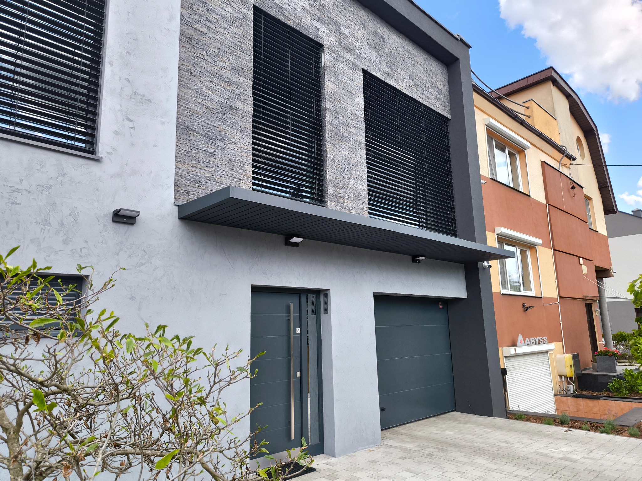 Nowoczesna elewacja domu z szarego kamienia i grafitową stolarką okienną, garażem i drzwiami wejściowymi. Widok z dołu, błękitne niebo w tle.