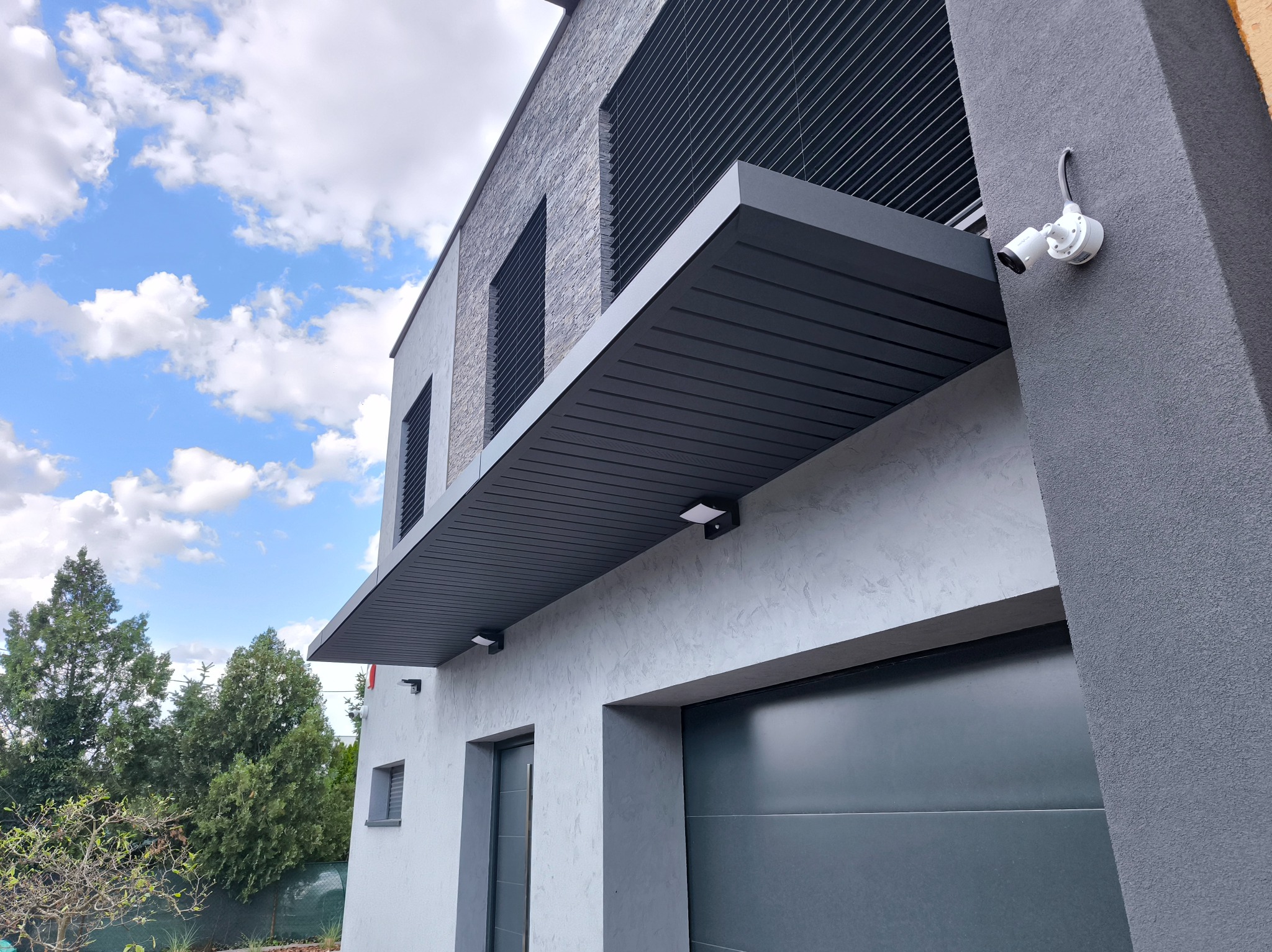 Nowoczesna, szara pergola metalowa nad wejściem do domu z kamerą, widziana z dołu pod kątem. Elementy dekoracyjne z kamienia. Błękitne niebo z chmurami w tle.