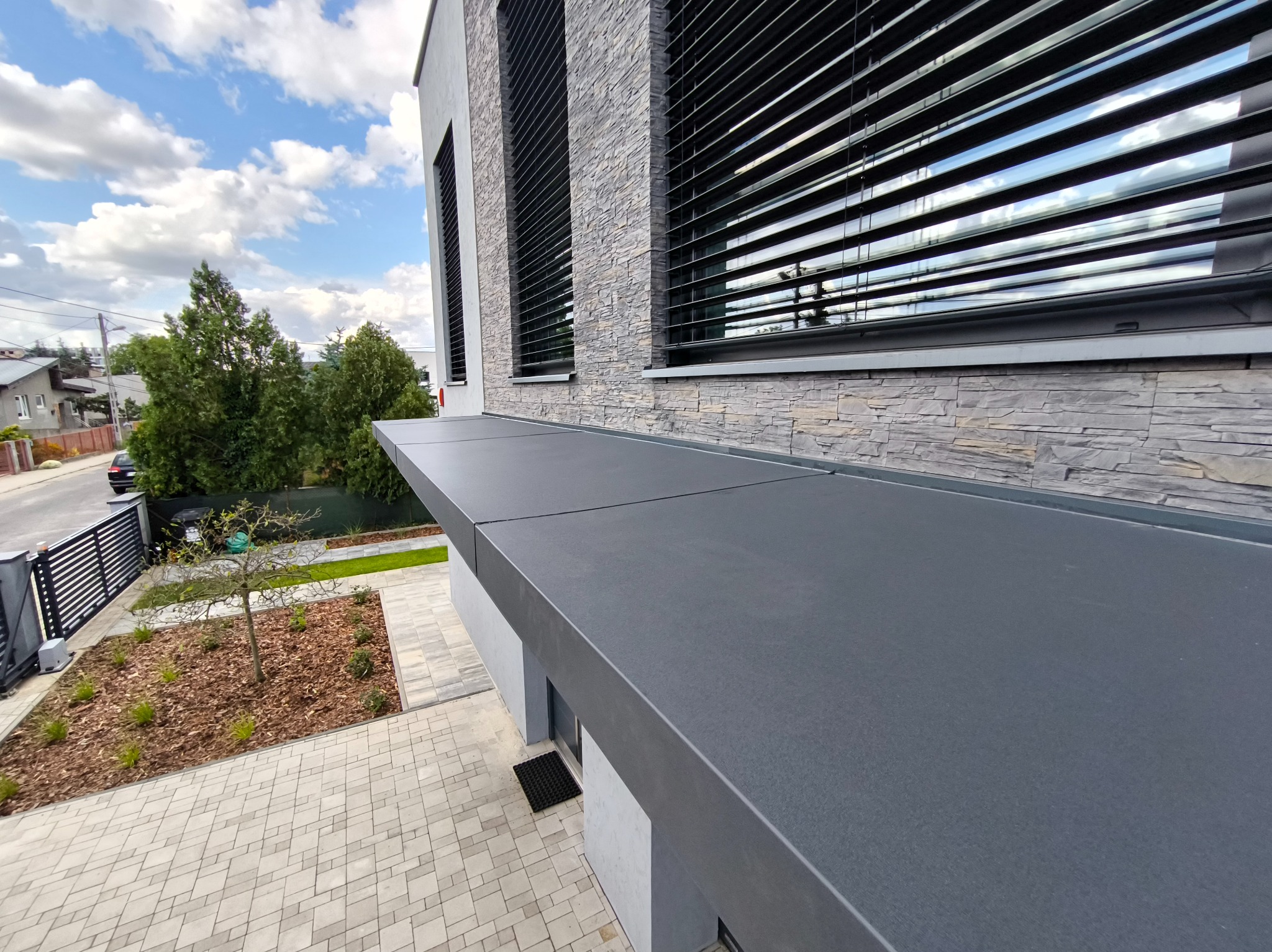 Nowoczesna pergola w kolorze antracytu, przylegająca do budynku z kamienną elewacją i żaluzjami. Widok z góry na kostkę brukową i fragment ogrodu.