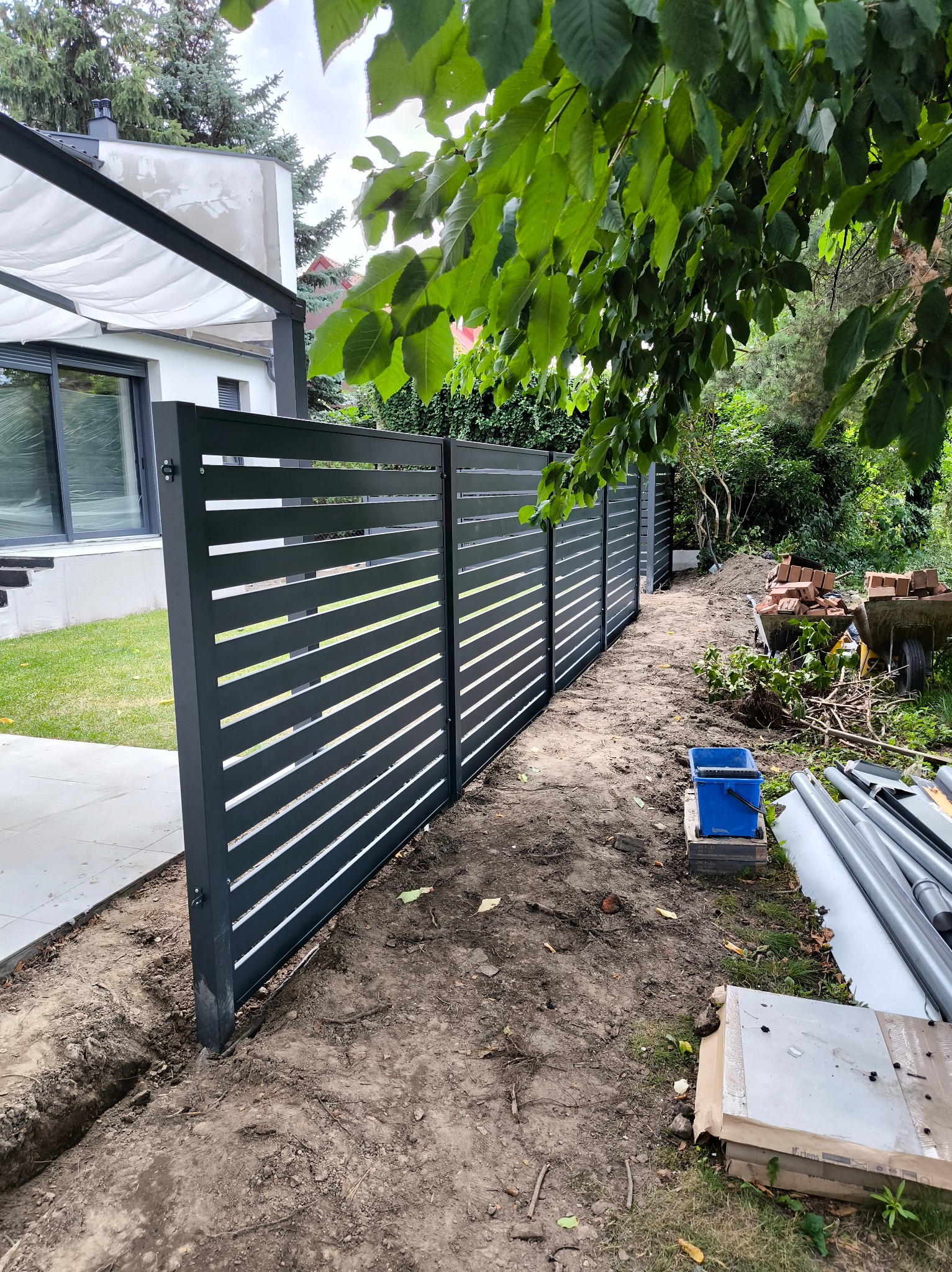 Nowoczesne ogrodzenie panelowe z poziomymi szczeblami, świeżo zamontowane, z widocznymi narzędziami i elementami montażowymi w tle. Dom z pergolą w tle.