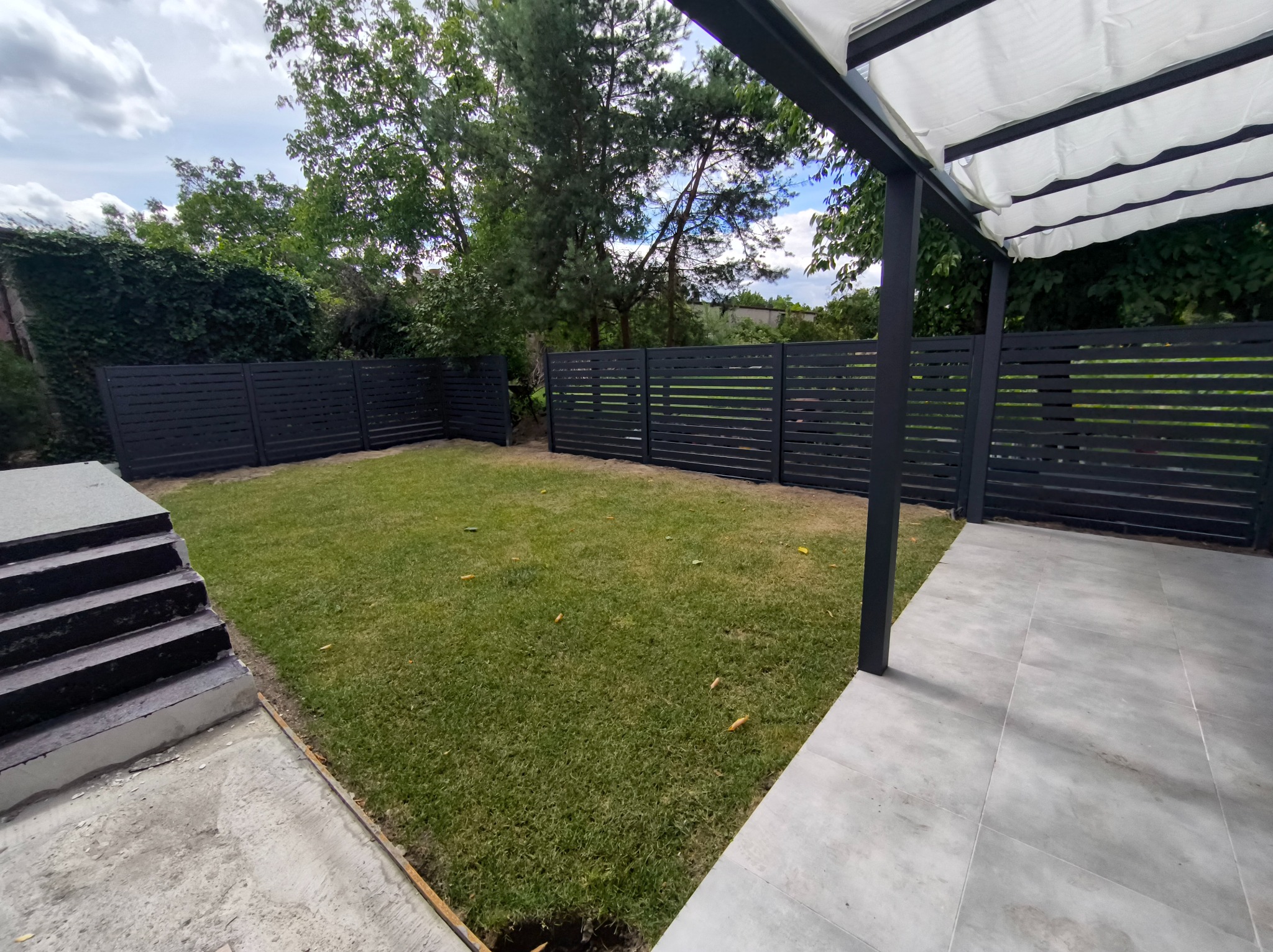 Nowoczesne, poziome ogrodzenie panelowe w kolorze antracytu otacza zadbany trawnik. Widoczna pergola z jasnym zadaszeniem i betonowe schody. Całość w otoczeniu zieleni.