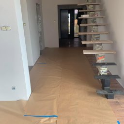 M&P Solid - Wnętrze w trakcie remontu: zabezpieczona podłoga brązowym papierem, widoczne schody wspornikowe z drewnianymi stopniami i metalowymi mocowaniami, ściany pomalowane na szaro.