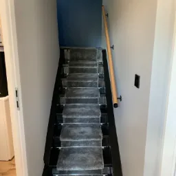 Schody z czarnymi stopniami pokryte szarym dywanem, drewniana poręcz zamontowana na białej ścianie, niebieska ściana w tle.