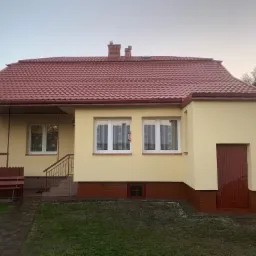 Dom jednorodzinny z nową, czerwoną dachówką i świeżo pomalowaną elewacją w odcieniach beżu, widoczny z perspektywy trawnika z kostką brukową. W tle błękitny van.