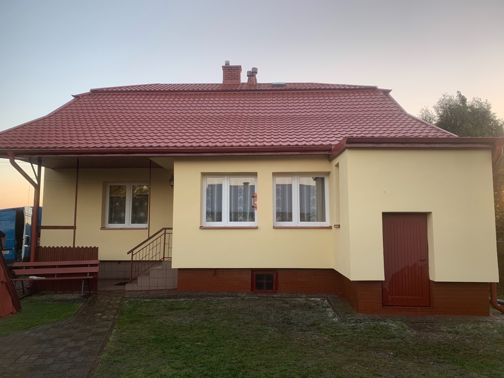 Dom jednorodzinny z nową, czerwoną dachówką i świeżo pomalowaną elewacją w odcieniach beżu, widoczny z perspektywy trawnika z kostką brukową. W tle błękitny van.