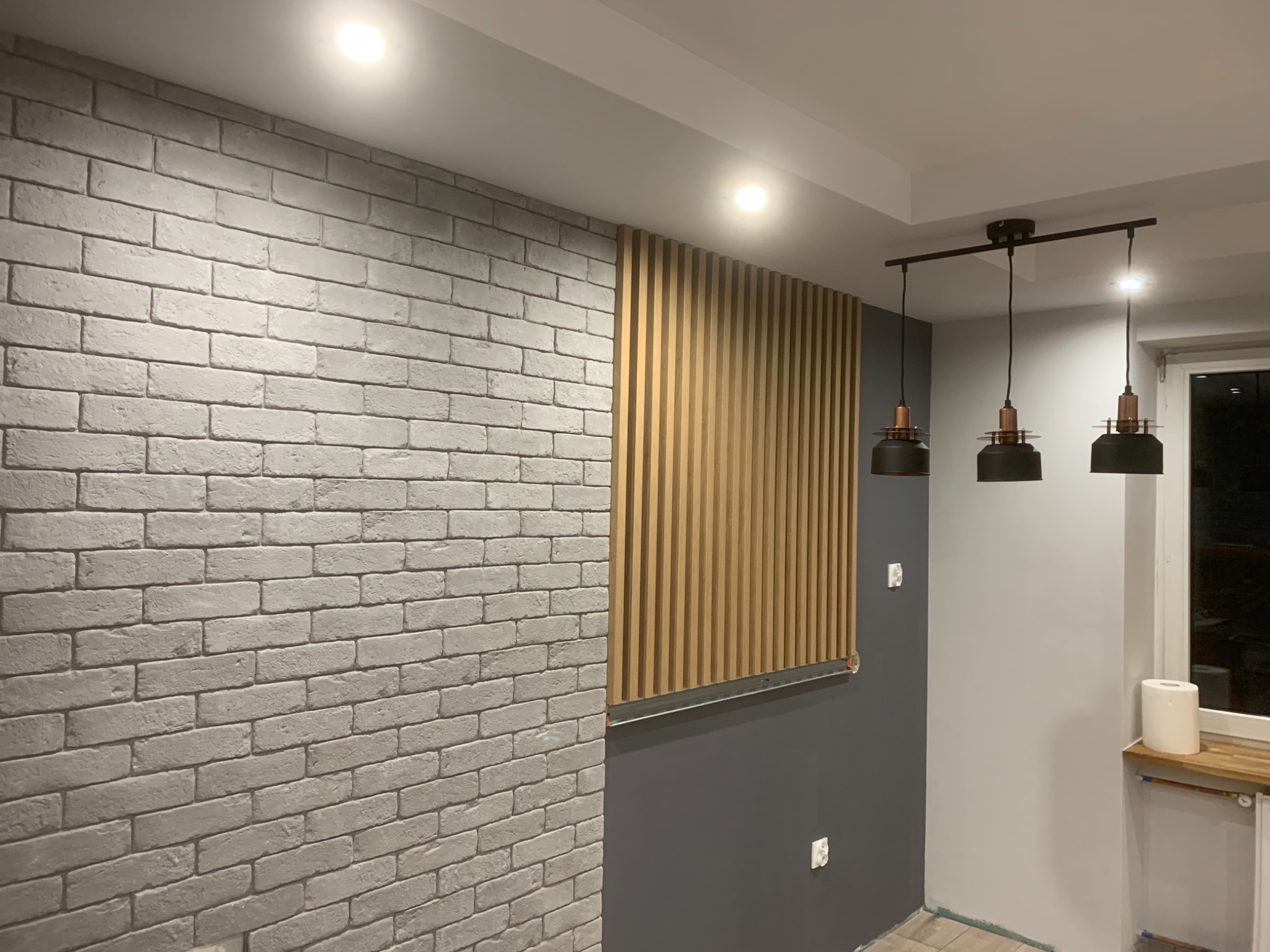 Wnętrze pokoju z szarą ceglaną ścianą, drewnianymi lamelami, szarą ścianą, trzema czarnymi lampami wiszącymi z miedzianymi akcentami i oświetleniem punktowym na suficie.
