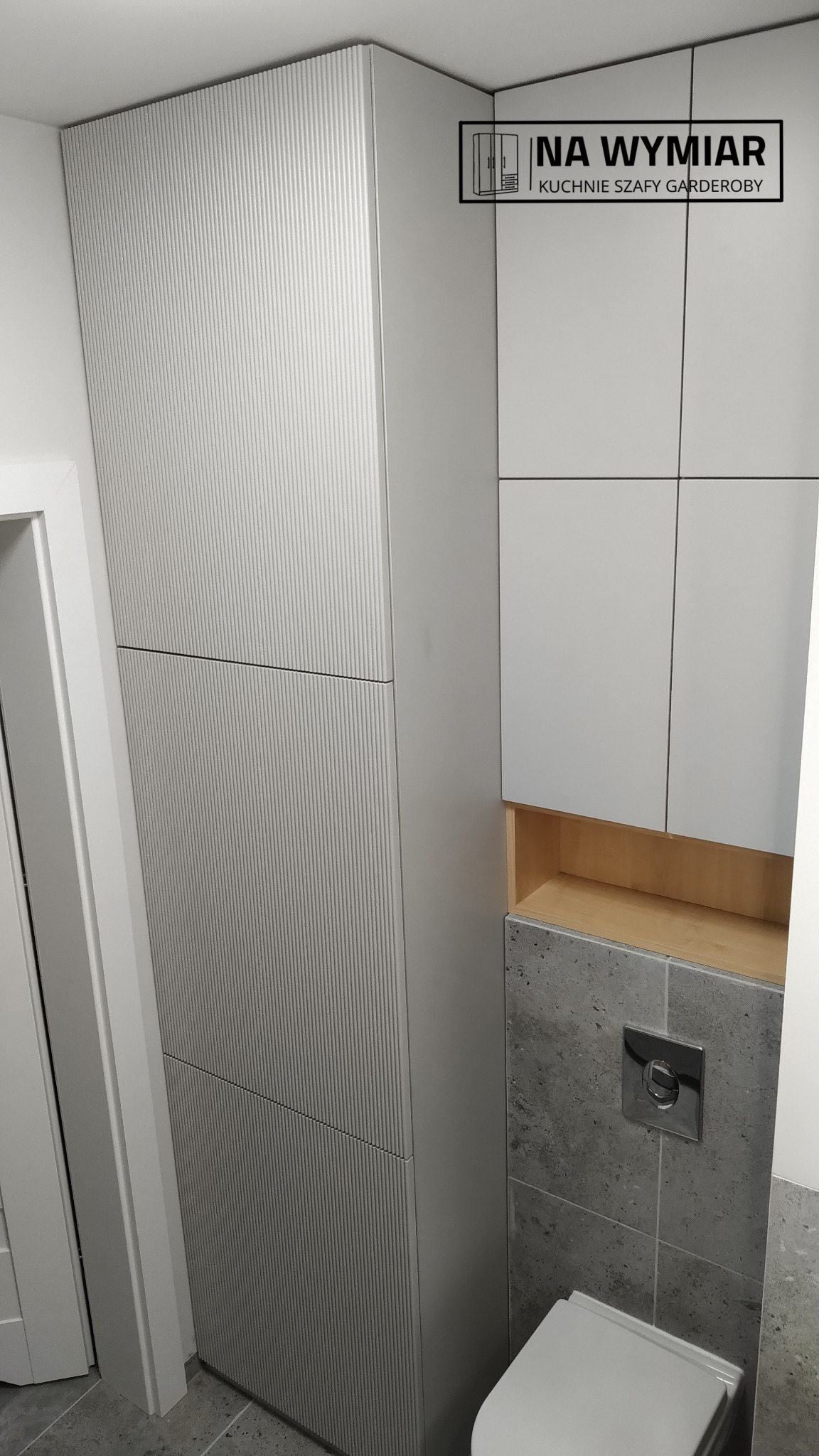 Wysoka, szara szafa wnękowa z ryflowanymi frontami w nowoczesnej łazience, widoczny fragment toalety i logo firmy na szafce obok.