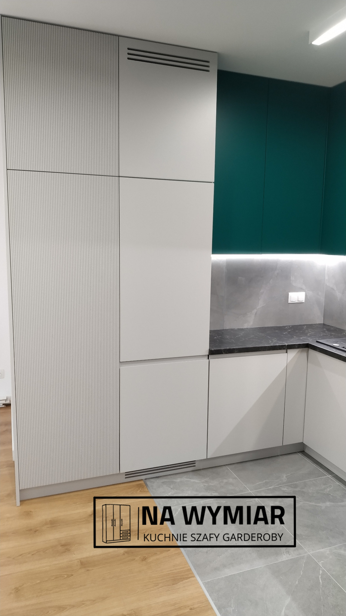 Nowoczesna zabudowa kuchenna: białe szafki z frezowanymi frontami, ciemnozielone szafki wiszące, szary blat i płytki imitujące marmur, widoczny fragment drewnianej podłogi.