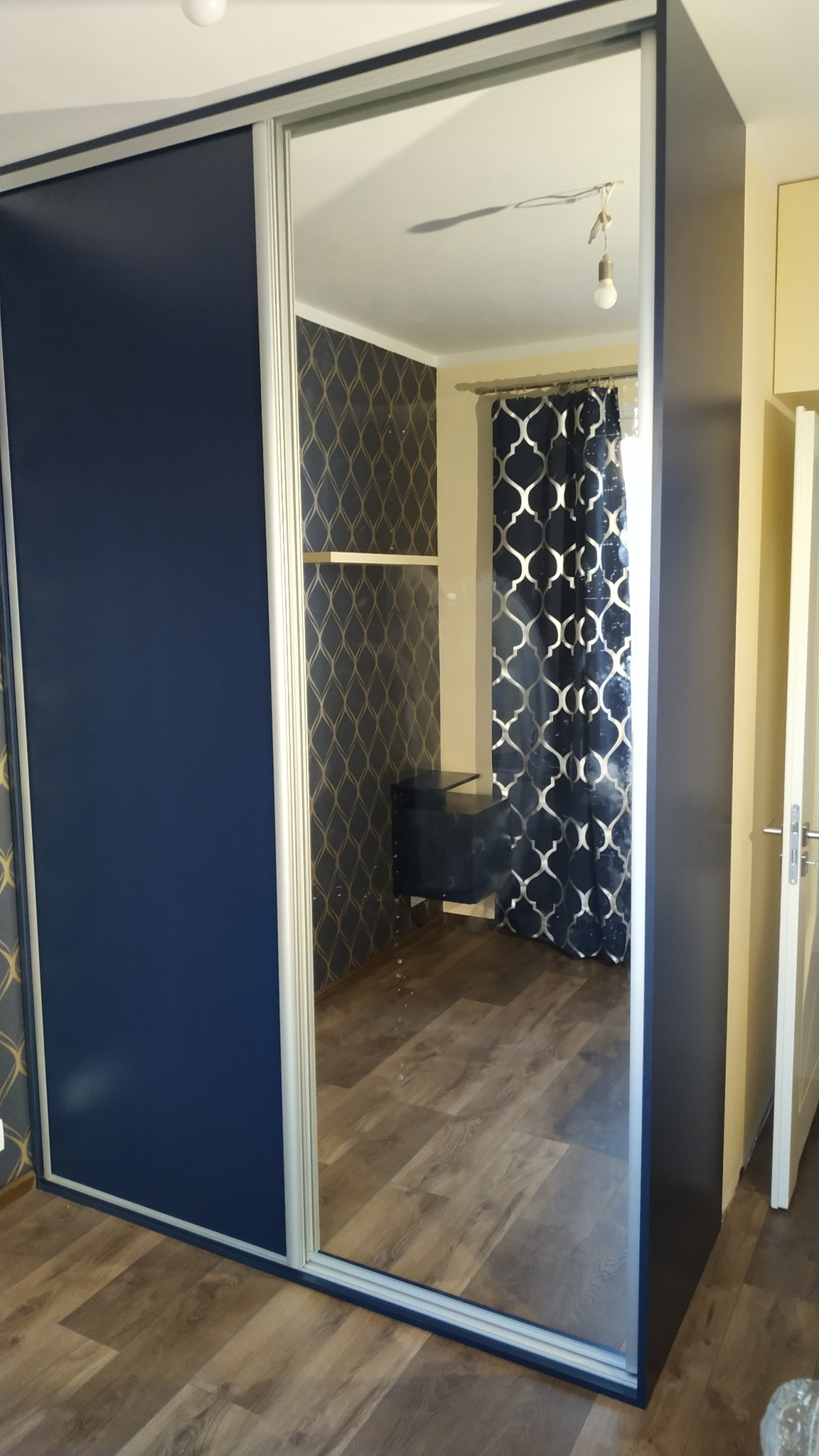 Szafa wnękowa z przesuwnymi drzwiami w kolorze granatowym, z lustrem odbijającym wnętrze pokoju z ciemną tapetą w złote wzory i zasłonami w geometryczne wzory.