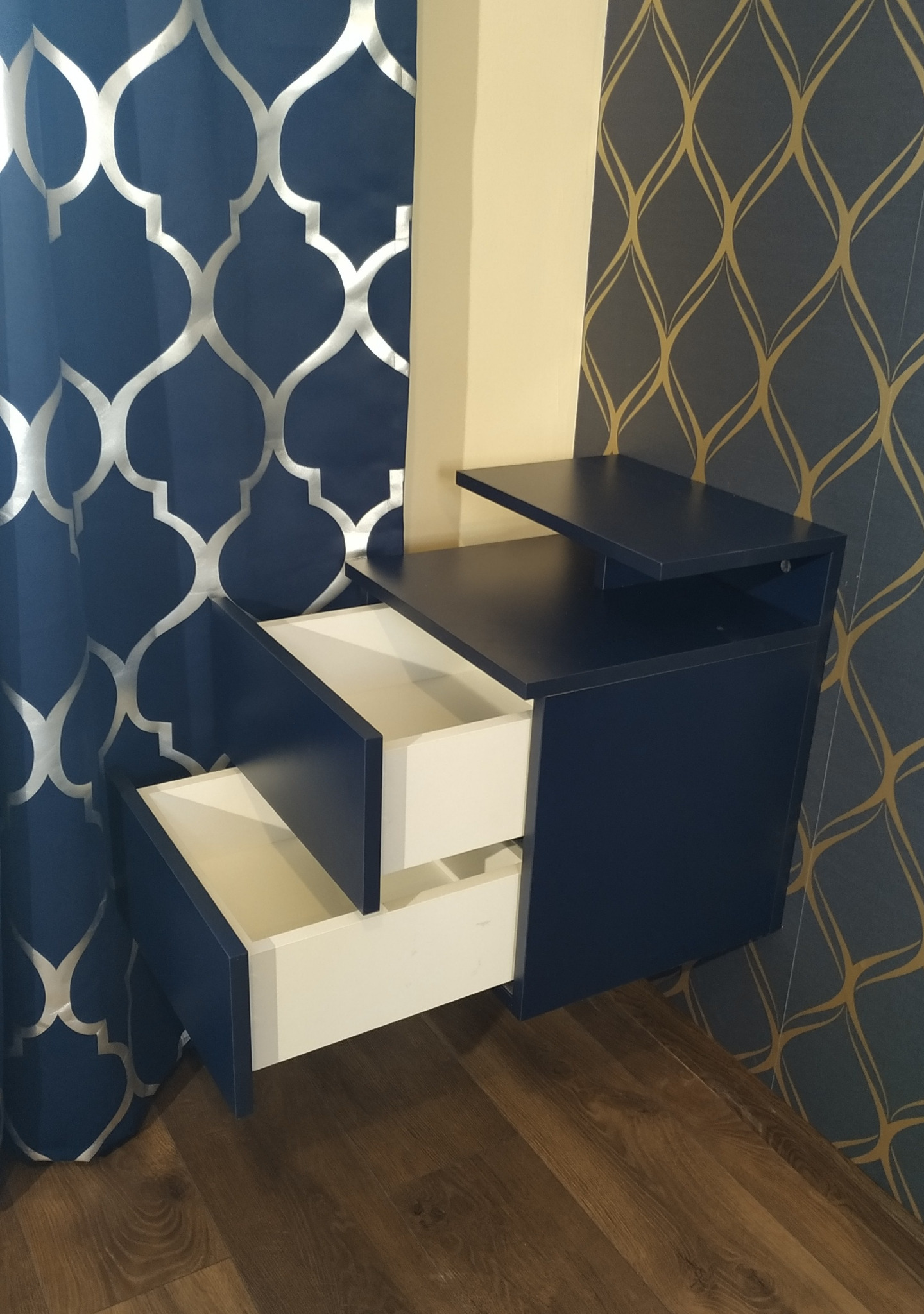 Niebieska szafka nocna z otwartymi białymi szufladami, zamontowana na ścianie z wzorzystą tapetą i zasłoną w tle.