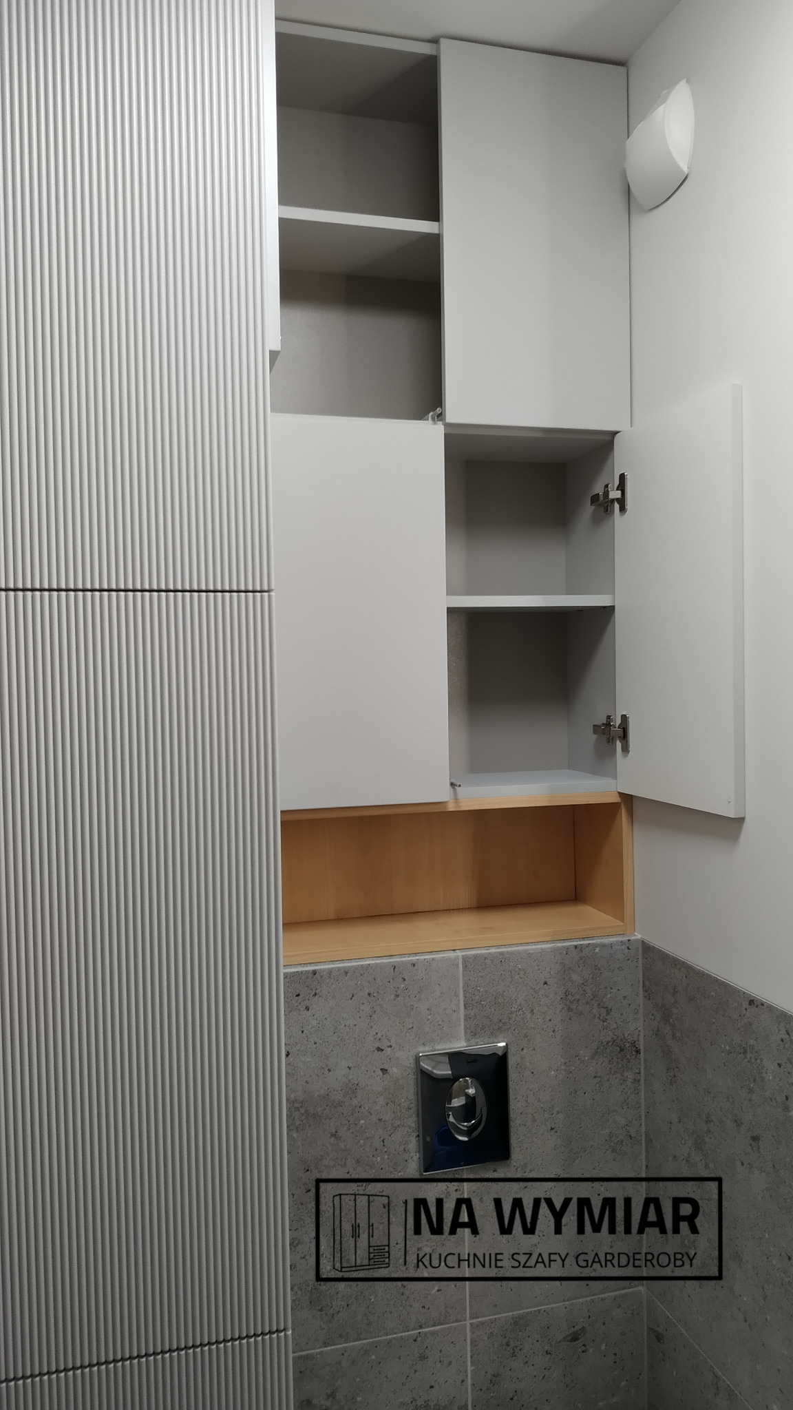 Szara szafka łazienkowa na wymiar z ryflowanym frontem, otwarte półki z drewnianym wykończeniem i logo firmy na tle płytek.