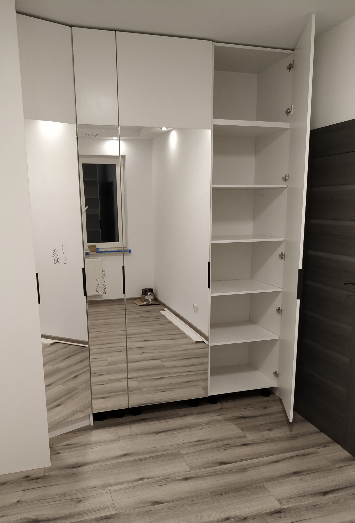 Biała szafa z lustrzanymi drzwiami wnękowymi, częściowo otwarta, ukazująca puste półki; widoczne notatki na lustrze i podłoga imitująca drewno.