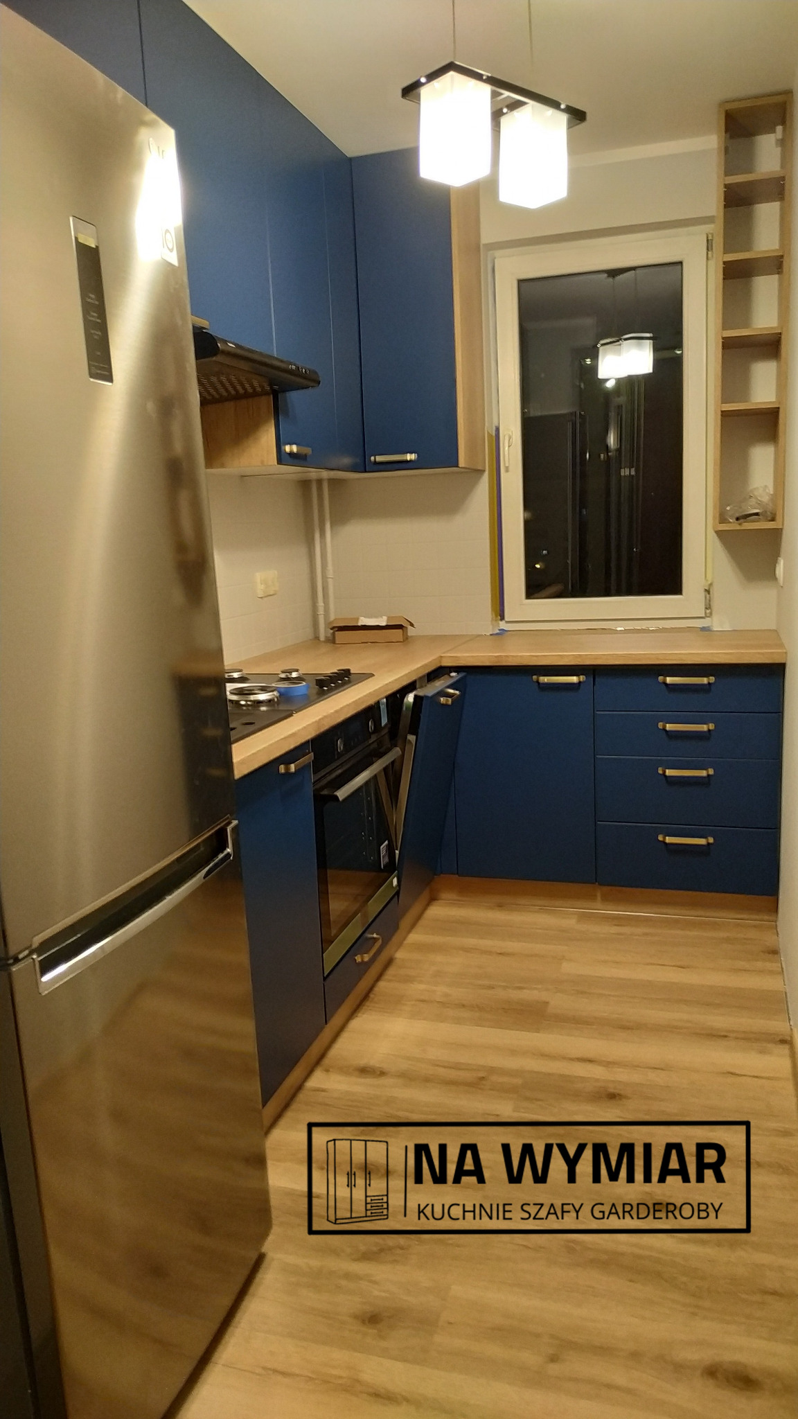 Niebieska zabudowa kuchenna z drewnianym blatem, widoczna lodówka ze stali nierdzewnej, płyta grzewcza i piekarnik pod oknem, drewniana podłoga.
