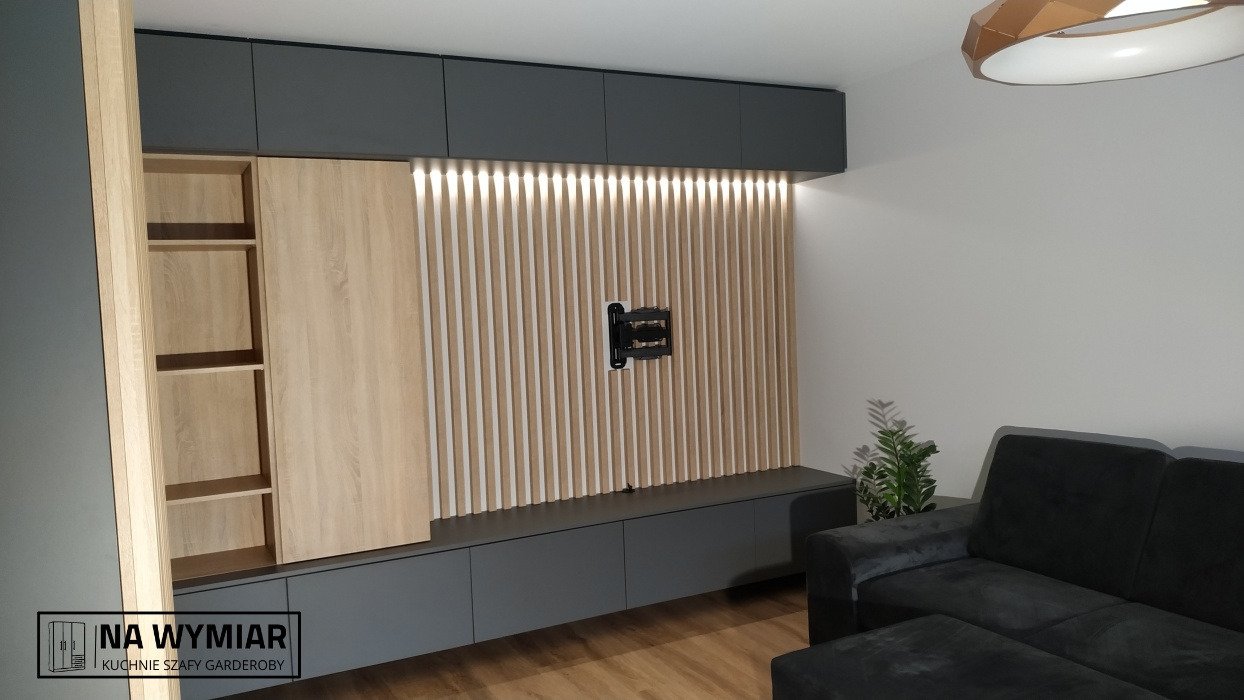 Nowoczesna zabudowa ściany telewizyjnej z drewnianymi lamelami, szarymi szafkami i otwartymi półkami z jasnego drewna, uzupełniona o czarną kanapę i roślinę doniczkową.
