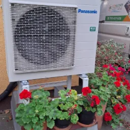 Biała jednostka zewnętrzna klimatyzacji Panasonic, zamontowana na szarej betonowej podstawie z doniczkami czerwonych pelargonii, na tle beżowej ściany budynku i białego samochodu dostawczego...