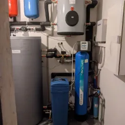 Kompletna kotłownia z dwoma zbiornikami ciśnieniowymi (czerwony i niebieski), dużym szarym zasobnikiem wody, filtrem wody z niebieską obudową oraz elementami instalacji hydraulicznej.