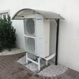 Zewnętrzna jednostka pompy ciepła Panasonic, zamontowana na metalowym stelażu i osłonięta półprzezroczystym daszkiem, otoczona drobnymi kamieniami na tle jasnej ściany budynku i kostki brukowej.