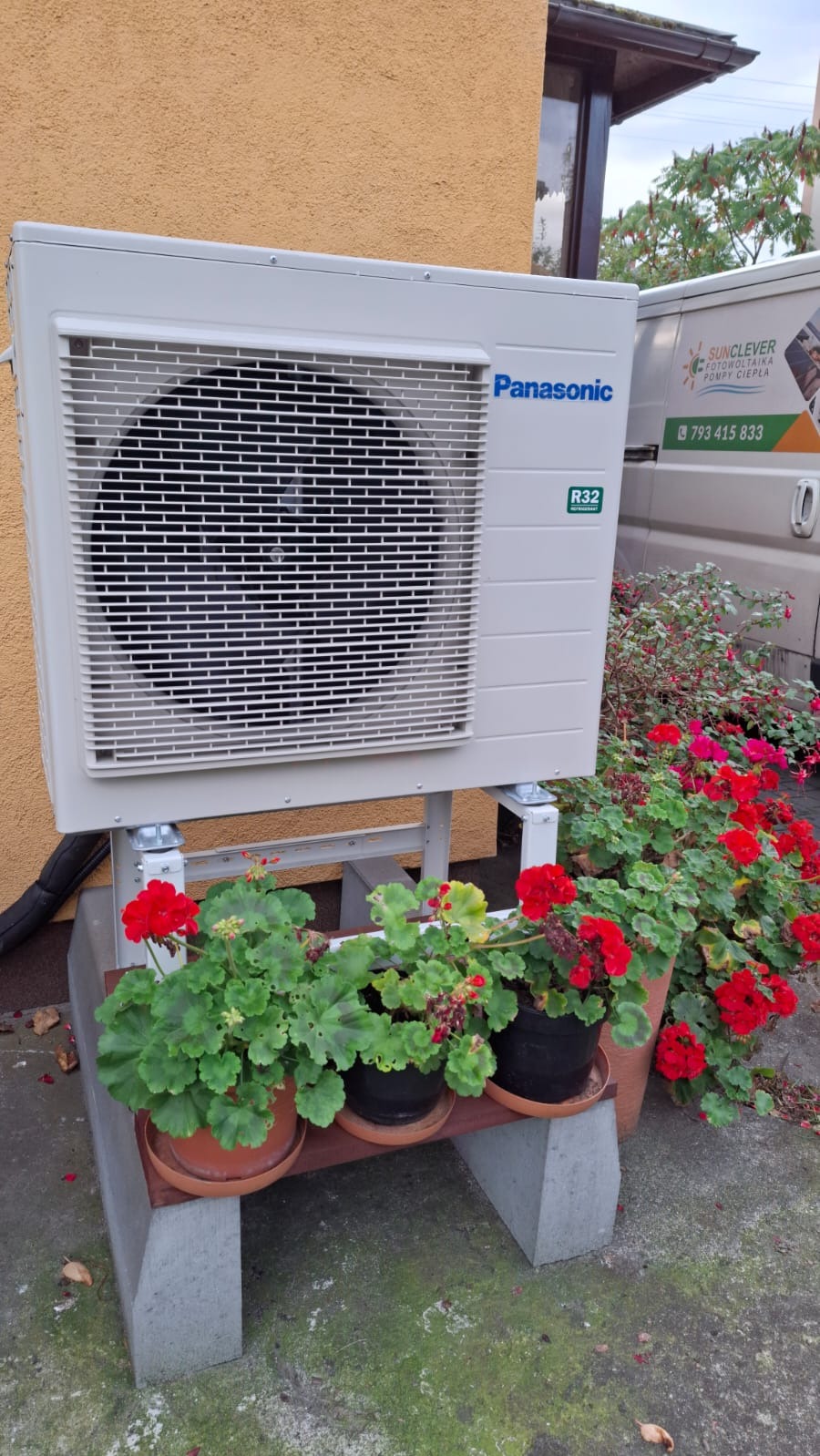 Biała jednostka zewnętrzna klimatyzacji Panasonic, zamontowana na szarej betonowej podstawie z doniczkami czerwonych pelargonii, na tle beżowej ściany budynku i białego samochodu dostawczego...