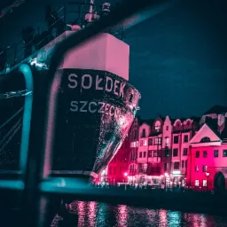 Nocne ujęcie dziobu statku 'Sołdek' w Szczecinie, z odbiciem świateł w wodzie i budynkami w tle, wszystko utrzymane w neonowej kolorystyce.