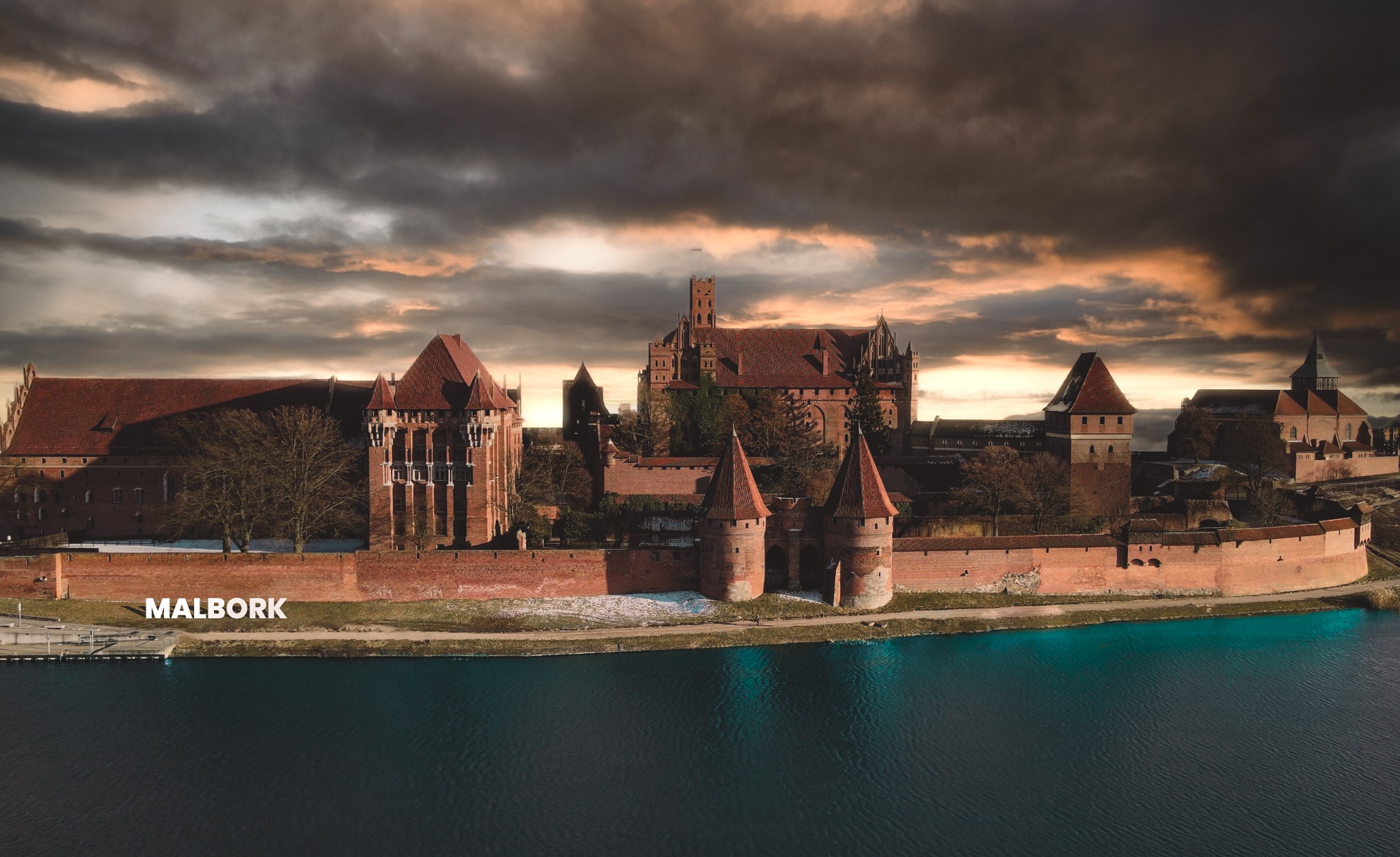 Panorama zamku w Malborku z lotu ptaka, widok na ceglane mury i wieże odbijające się w wodzie, na tle dramatycznego, zachmurzonego nieba oświetlonego promieniami zachodzącego słońca.