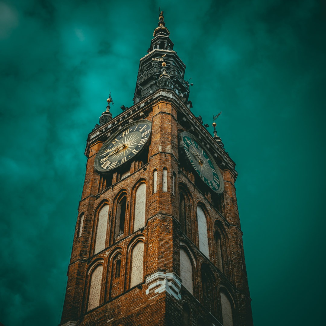 Wysoka, ceglana wieża zegarowa z dwoma tarczami zegarowymi, sfotografowana z dołu na tle ciemnego, burzowego nieba w odcieniach zieleni i szarości.