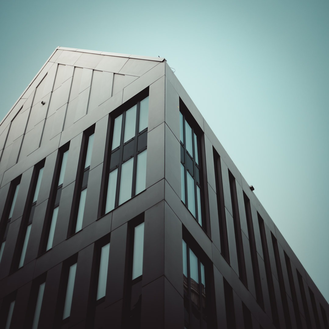 Perspektywiczny widok na nowoczesny biurowiec z ciemną elewacją i regularnym układem okien pod błękitnym niebem.