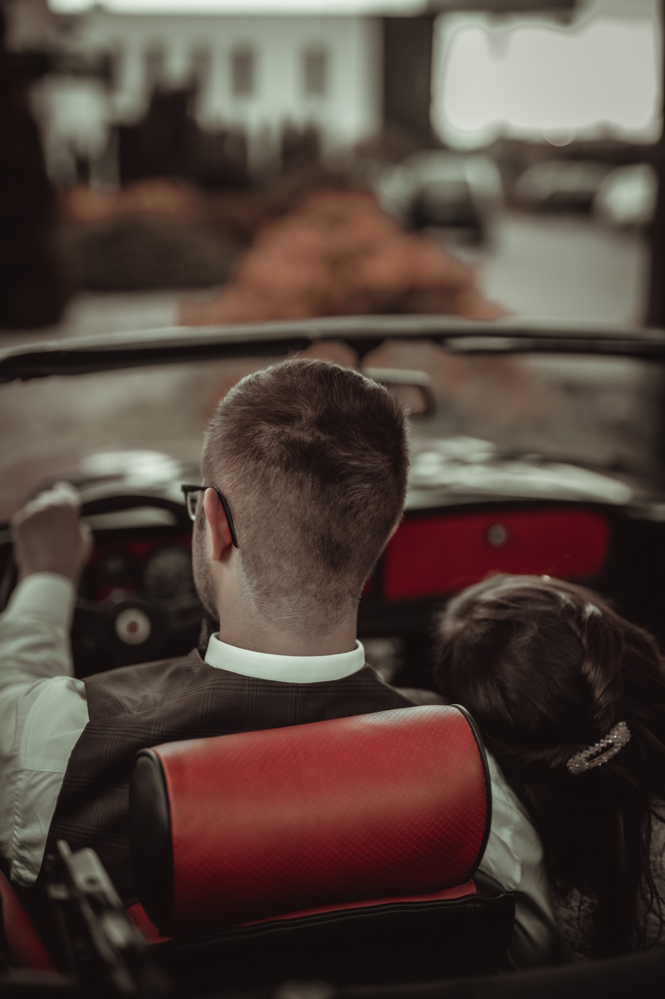 Para młoda w kabriolecie, widok z tyłu na kierowcę w okularach i pannę młodą z ozdobą we włosach, podczas sesji ślubnej w plenerze.