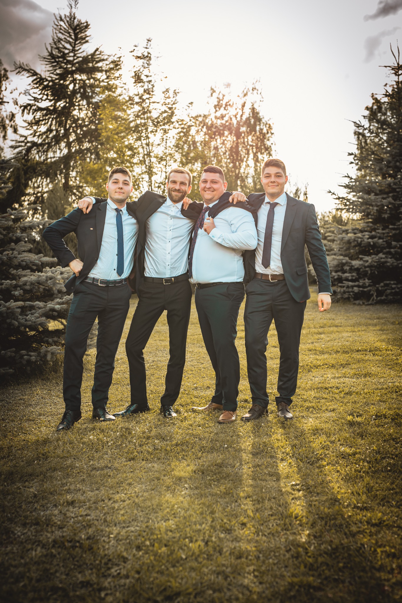 Czterech mężczyzn w garniturach, stojących obok siebie na trawie, z rękami na ramionach, w słoneczny dzień.