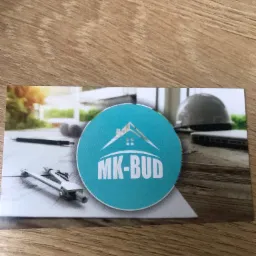 Wizytówka firmy MK-BUD z logo na tle rysunku architektonicznego, kasku budowlanego i laptopa, leżąca na drewnianym blacie.