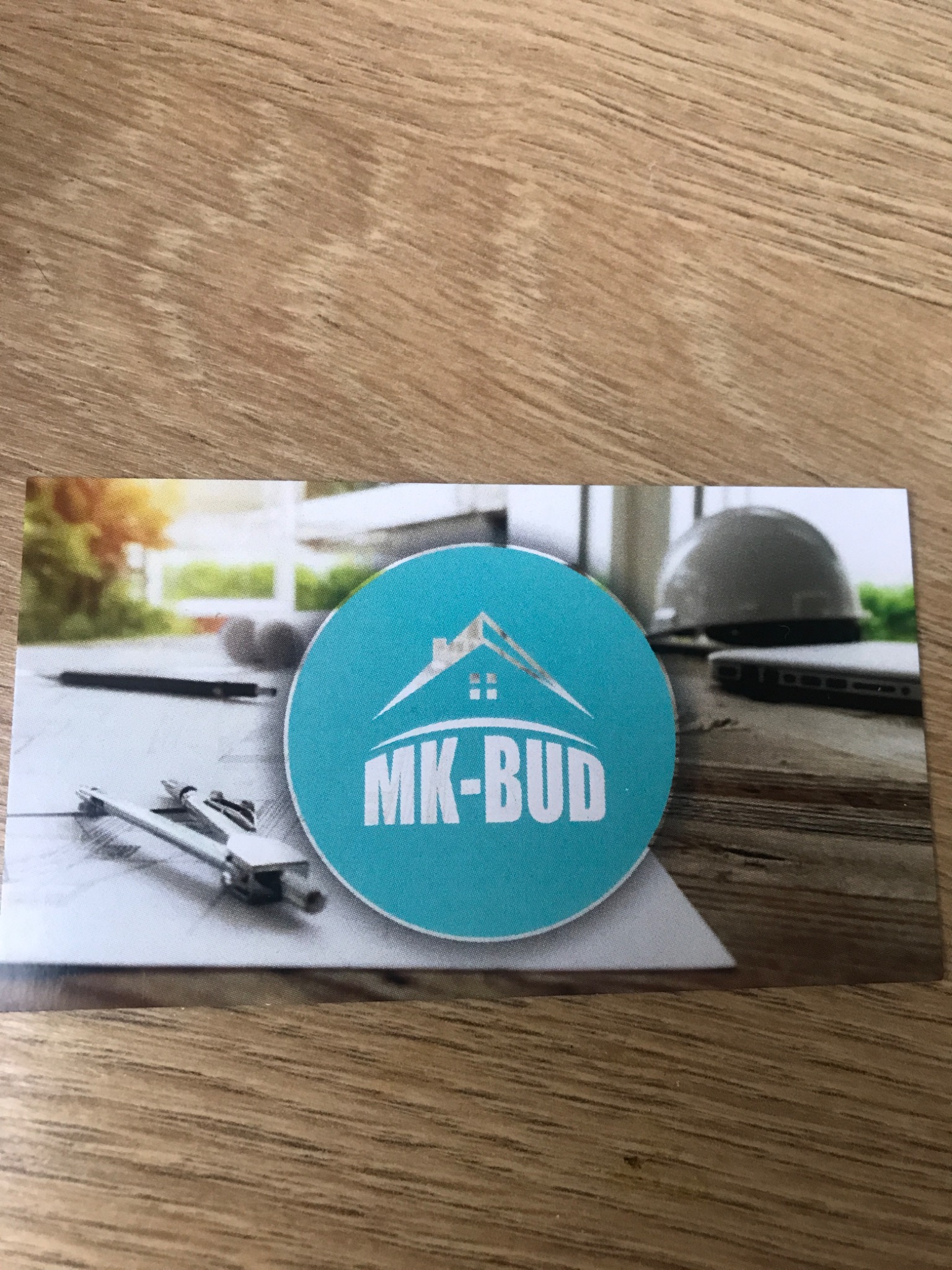 Wizytówka firmy MK-BUD z logo na tle rysunku architektonicznego, kasku budowlanego i laptopa, leżąca na drewnianym blacie.
